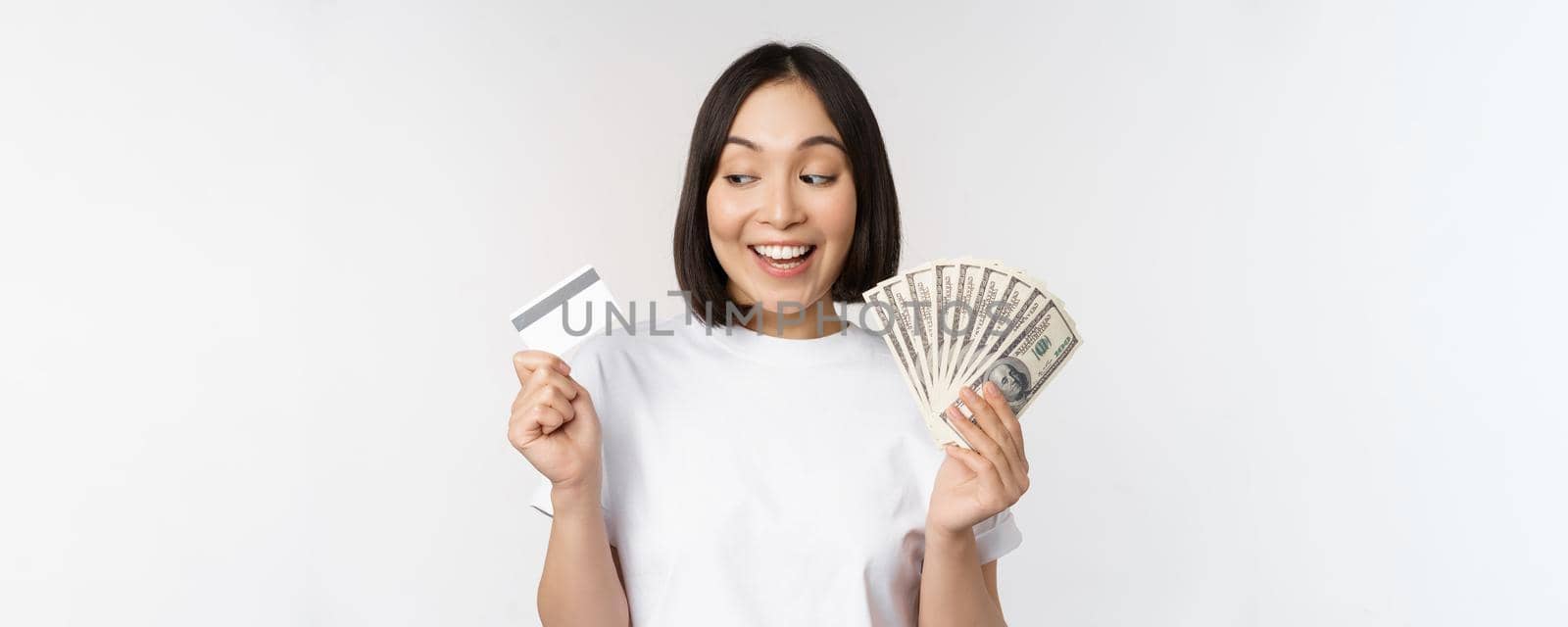 Portrait of asian woman smiling, holding credit card and money cash, dollars, standing in tshirt over white background by Benzoix