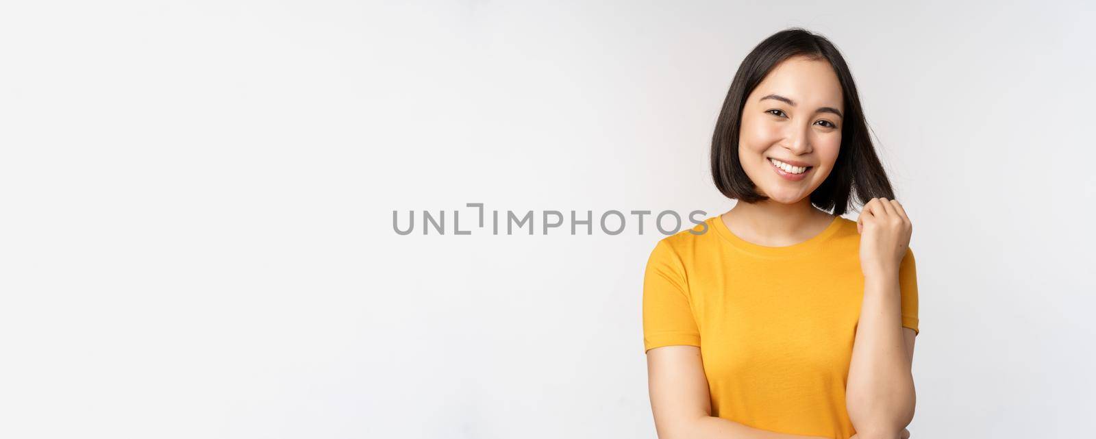 Beautiful romantic asian girl, smiling and playing with hair, looking happy at camera, standing in yellow t-shirt over white background by Benzoix