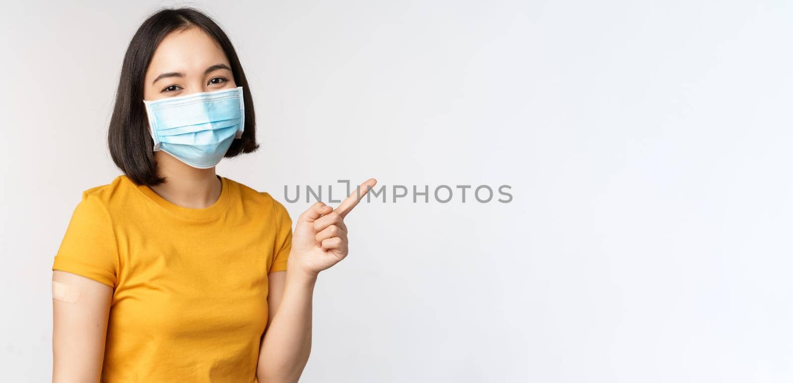 Covid-19, vaccination and healthcare concept. Portrait of cute asian girl in medical mask, has band aid on shoulder after coronavirus vaccine, standing over white background by Benzoix