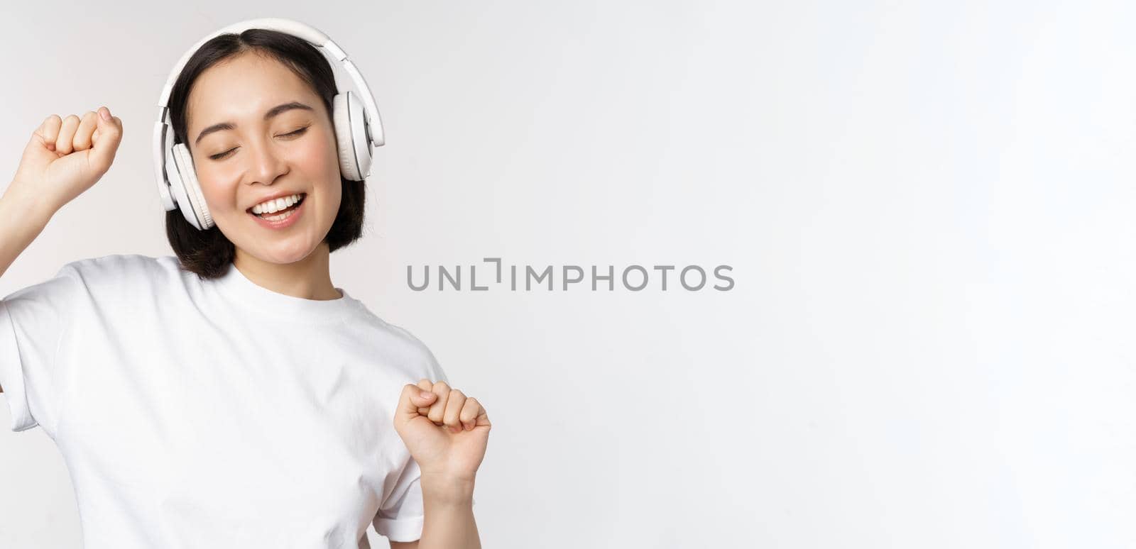 Dancing and singing asian woman, listening music in headphones, standing in earphones against white background by Benzoix