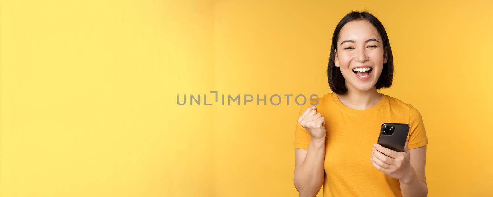 Joyful asian woman celebrating, holding mobile phone, winning, achieve goal on smartphone, standing over yellow background by Benzoix