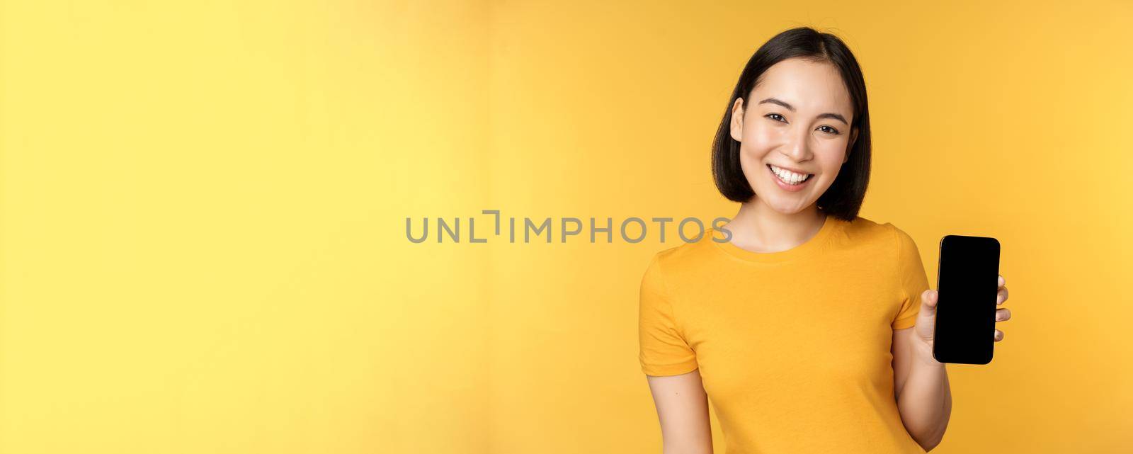 Beautiful happy asian girl showing mobile phone screen, application on smartphone gadget, standing over yellow background.