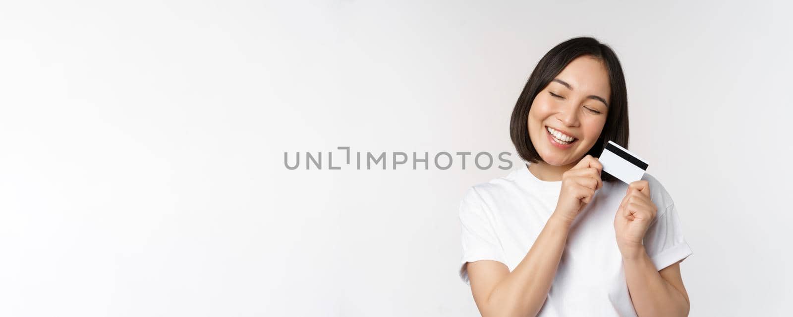 Image of smiling asian woman hugging credit card, buying contactless, standing in white tshirt over white background. Copy space