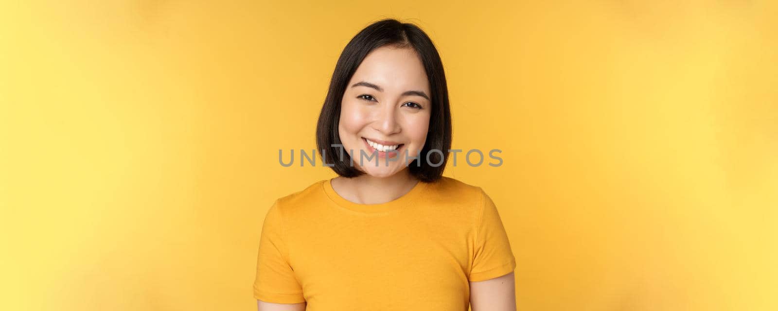 Close up portrait of beautiful asian woman smiling, looking cute and tender, standing against yellow background.