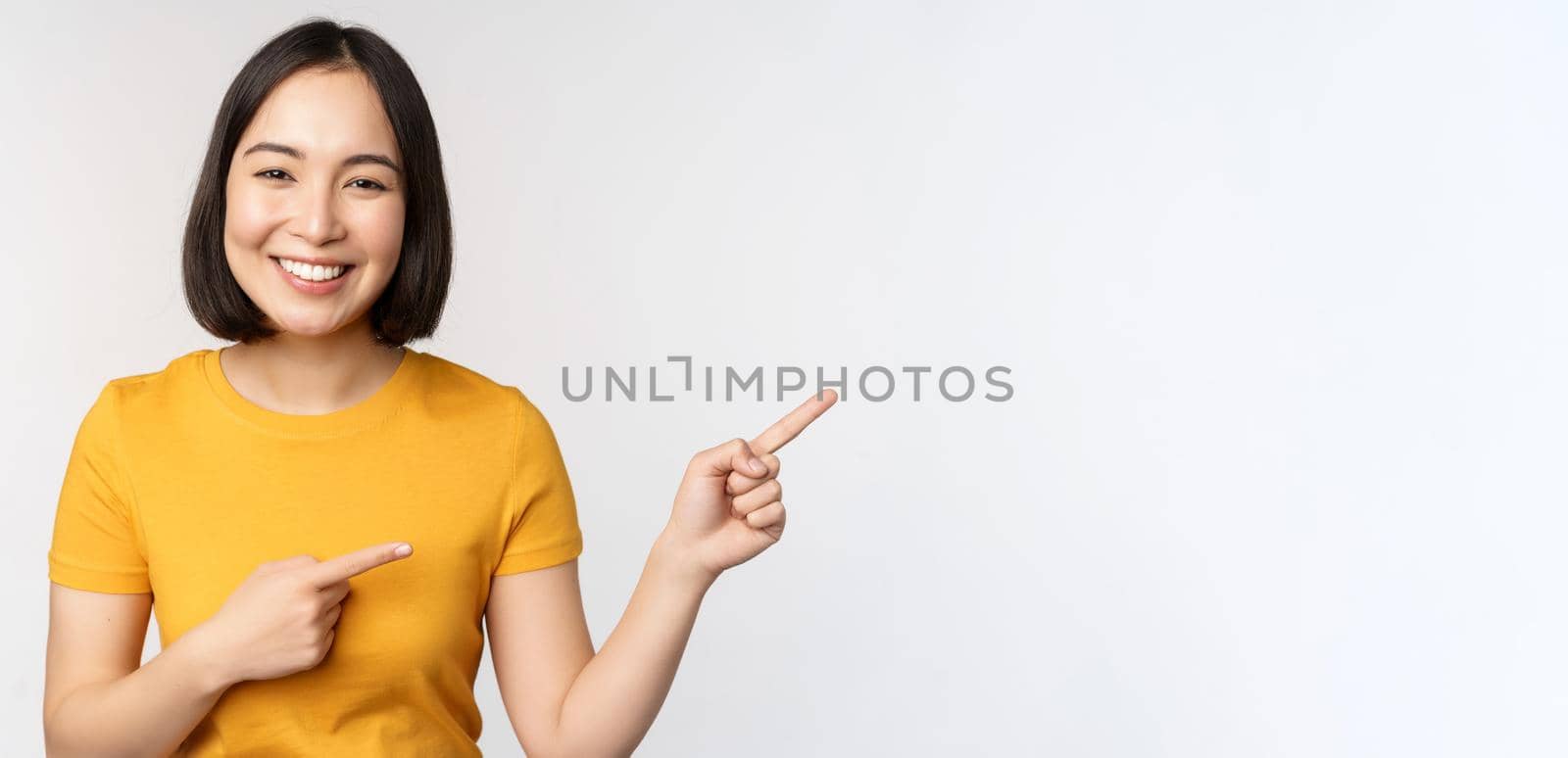 Amazed young asian woman, showing advertisement aside, pointing fingers right at promotion text, brand logo, standing happy against white background by Benzoix