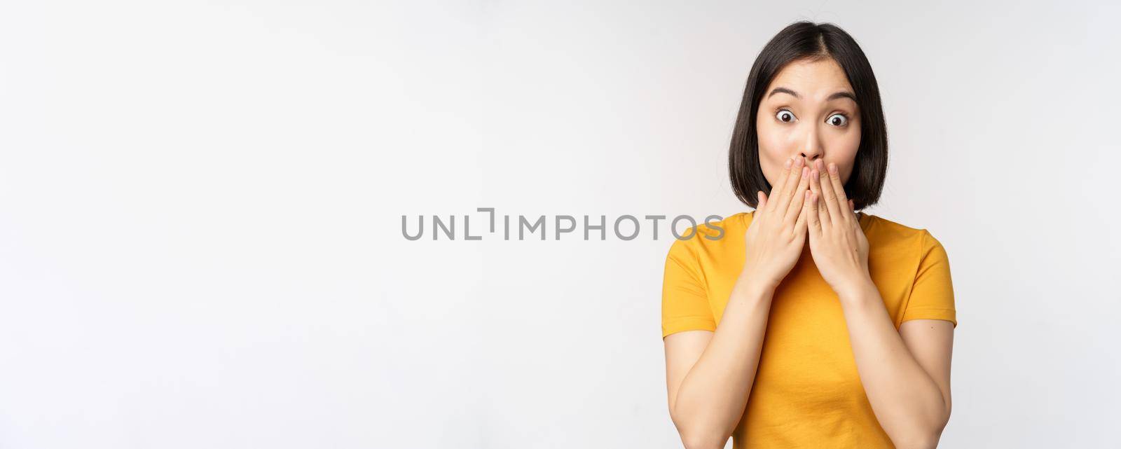 Surprised asian woman gasping, cover mouth with hands and looking amazed at camera, wearing yellow t-shirt, standing over white background.