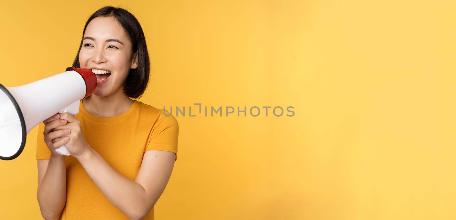 Announcement. Happy asian woman shouting loud at megaphone, recruiting, protesting with speaker in hands, standing over yellow background by Benzoix