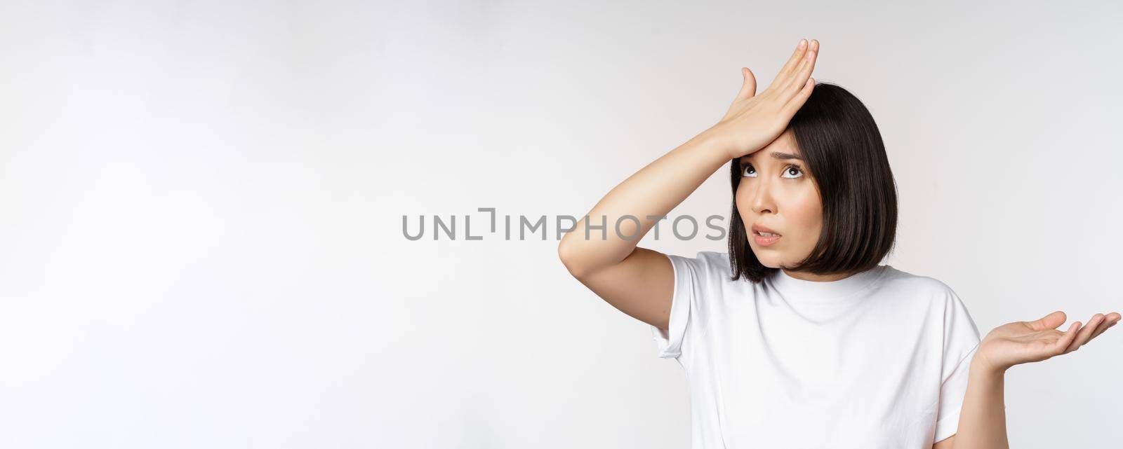 Annoyed korean girl facepalm, slap forehead and shrugging, confused by smth, standing over white background by Benzoix