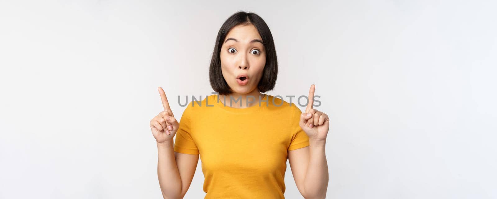 Portrait of impressed asian girl, pointing fingers up, say wow, looking amazed at camera, standing over white background. Copy space