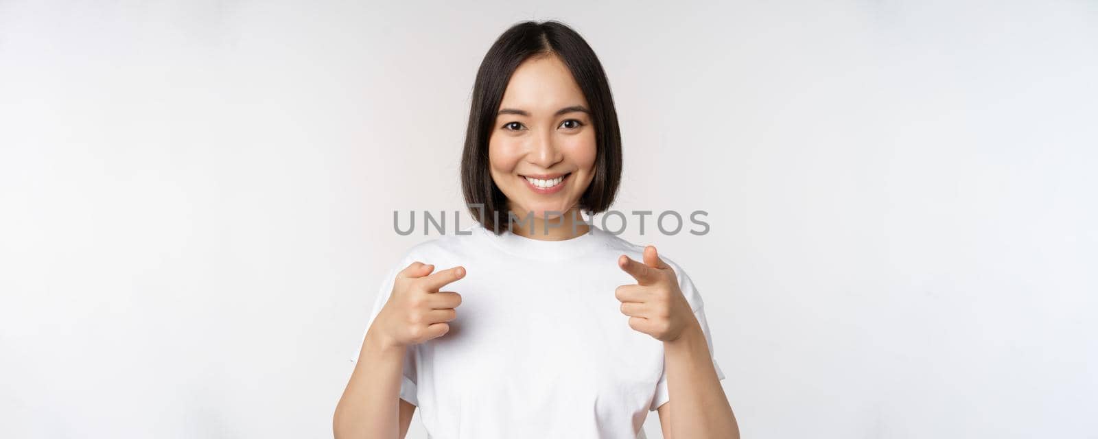 Image of smiling asian girl pointing fingers at camera, choosing, inviting you, congratulating, standing in tshirt over white background.