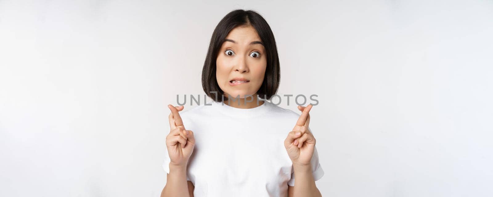 Hopeful asian girl cross fingers, making wish, anticipating, wishing smth, standing in tshirt over white background. Copy space