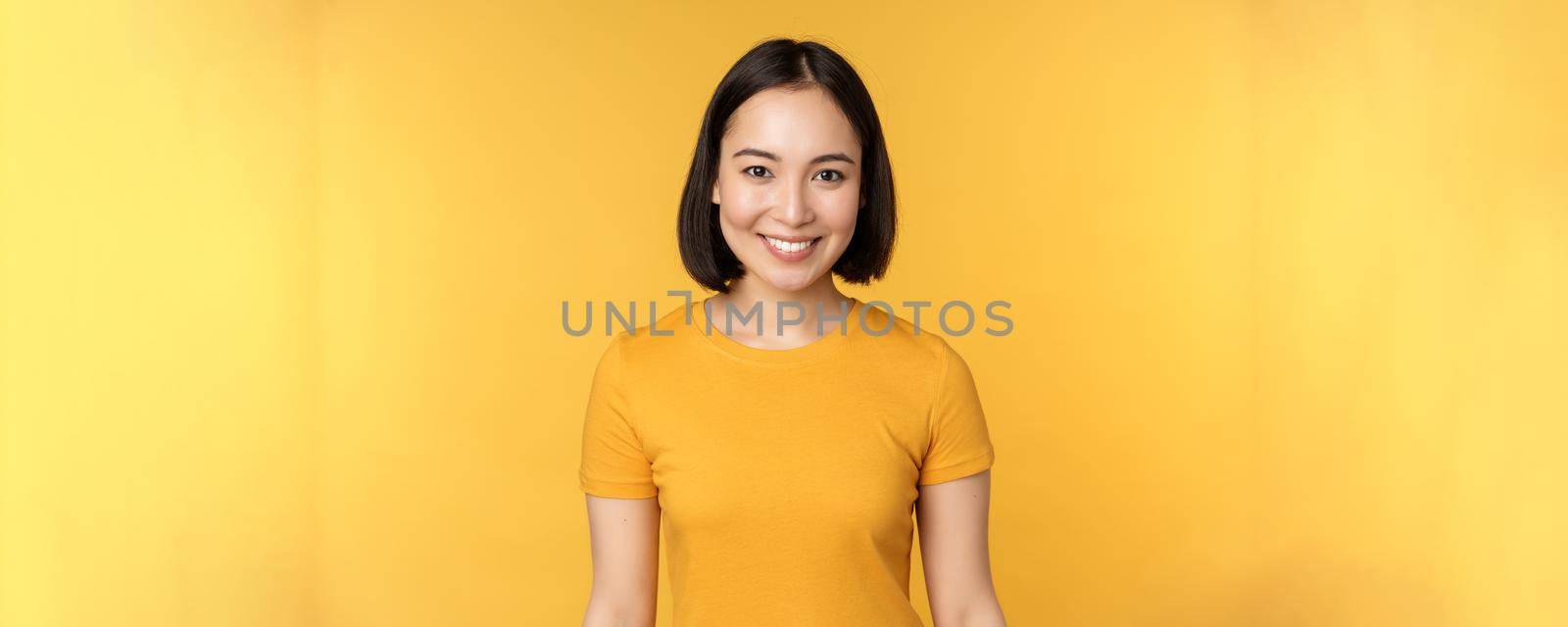 Portrait of young modern asian woman, smiling happy with white teeth, looking confident at camera, wearing casual t-shirt, standing over yellow background.