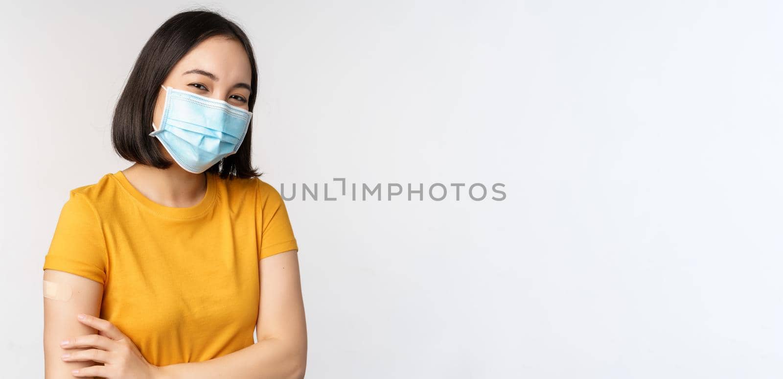 Covid-19, vaccination and healthcare concept. Cute asian girl in medical face mask, holding shoulder with band aid, got vaccine from coronavirus, smiling pleased, white background.