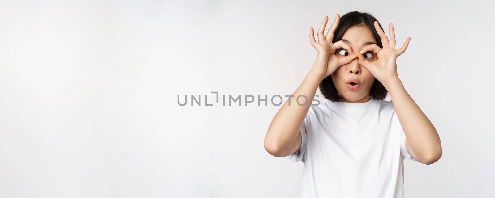 Funny young asian woman, korean girl making eyes glasses gesture, looking happy at camera, standing over white background by Benzoix