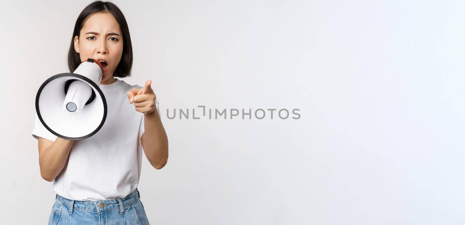 Angry asian woman with megaphone, scolding, accusing someone, protesting with speakerphone on protest, standing over white background by Benzoix