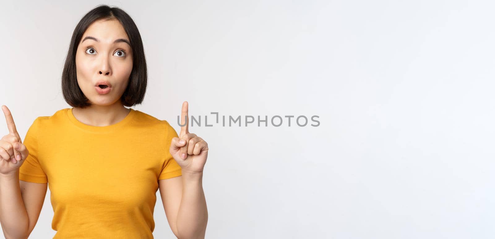 Happy beautiful asian girl showing advertisement, pointing fingers up, standing over white background by Benzoix