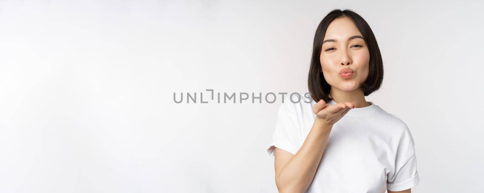 Lovely young asian woman, sending air kiss and looking coquettish at camera, standing in tshirt over white background.