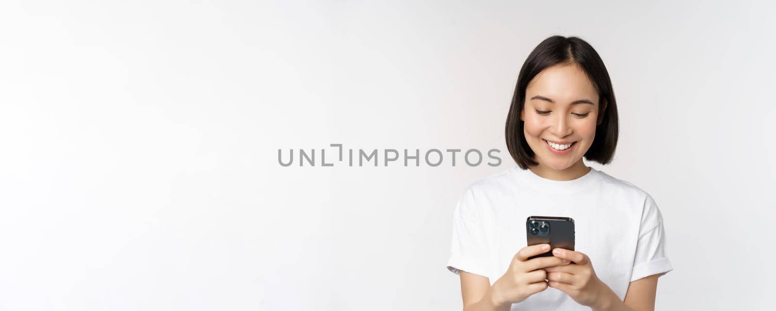 Portrait of smiling asian woman using mobile phone, chatting, texting message, standing in tshirt over white background. Copy space
