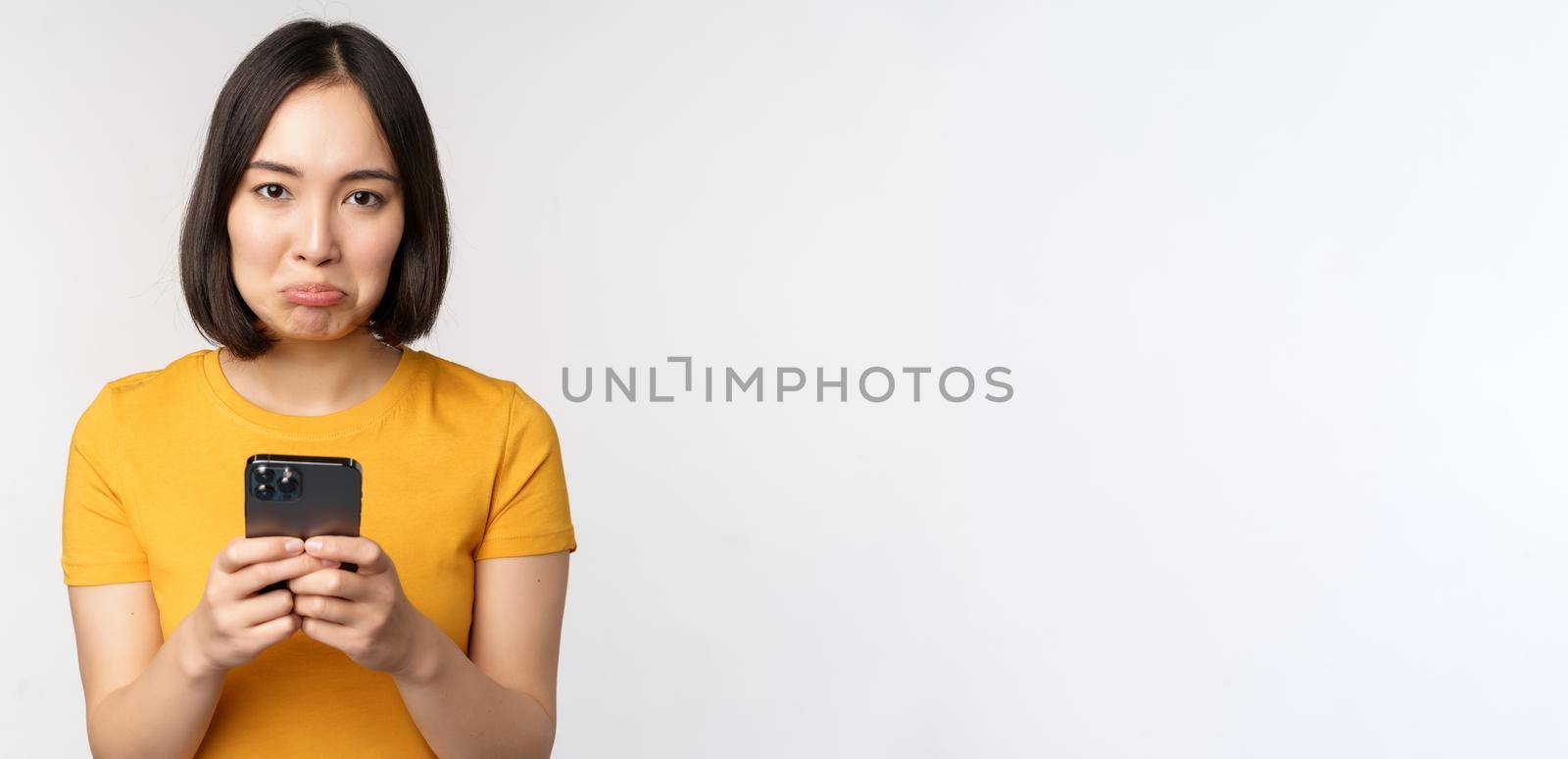 Sad asian woman holding smartphone, looking upset with regret, standing in yellow tshirt against white background by Benzoix