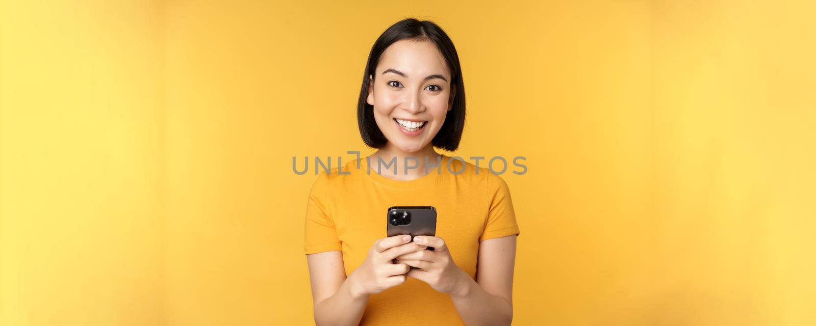 Technology. Smiling asian woman using mobile phone, holding smartphone in hands, standing in t-shirt against yellow background by Benzoix