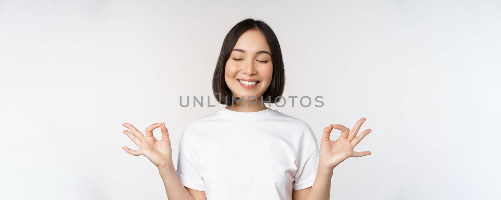 Portrait of young asian woman meditating, smiling pleased and practice yoga, close eyes and meditate, breathing calm, standing over white background by Benzoix