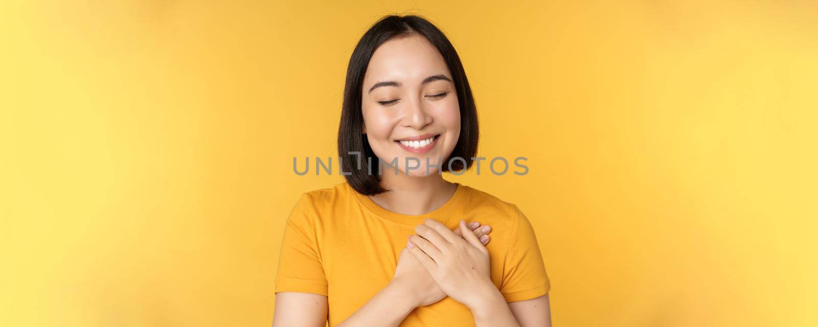 Beautiful asian woman, smiling with tenderness and care, holding hands on heart, standing in tshirt over yellow background.