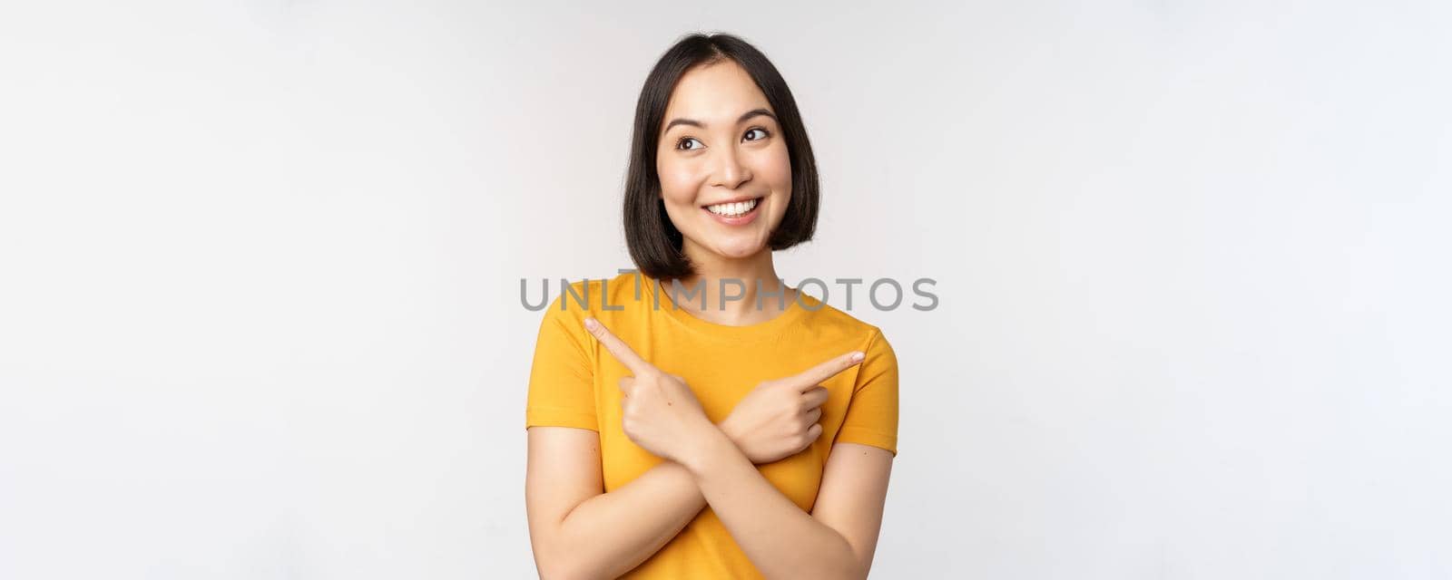 Cute asian girl pointing fingers sideways, showing left and right promo, two choices, variants of products, standing in yellow tshirt over white background.