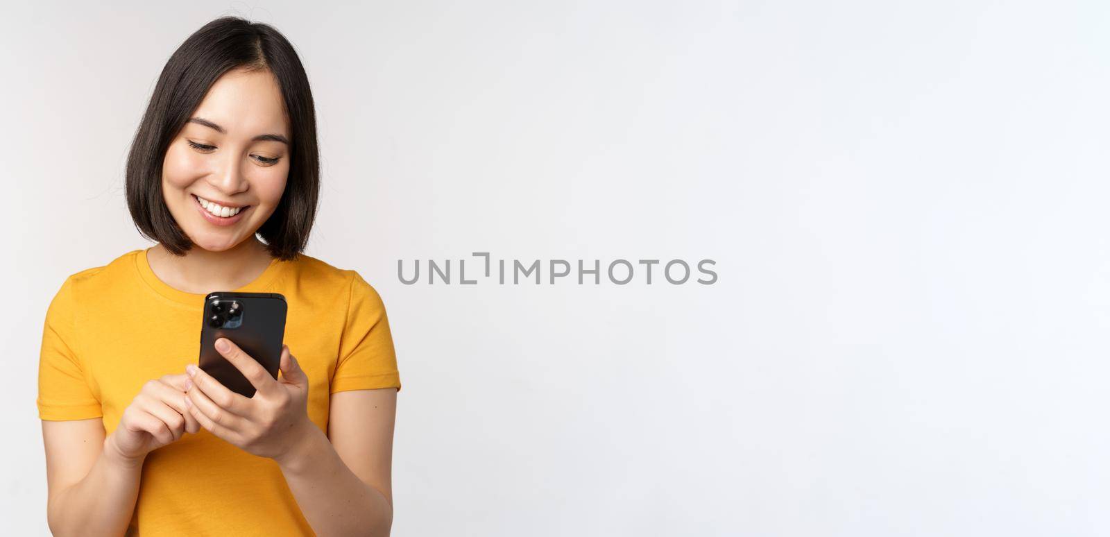 People and technology concept. Smiling asian girl using smartphone, texting on mobile phone, standing against white background by Benzoix