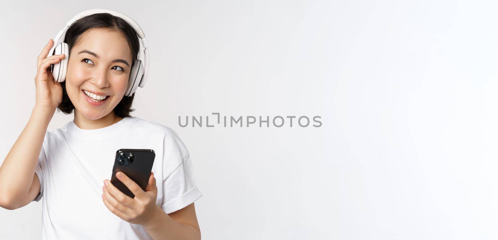 Beautiful modern asian girl, listening music in headphones, holding mobile phone, using smartphone app, standing over white background by Benzoix