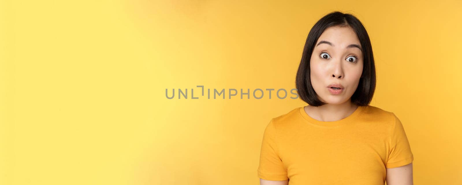 Close up portrait of asian girl showing surprised reaction, raising eyebrows amazed, reacting to big news, standing over yellow background.