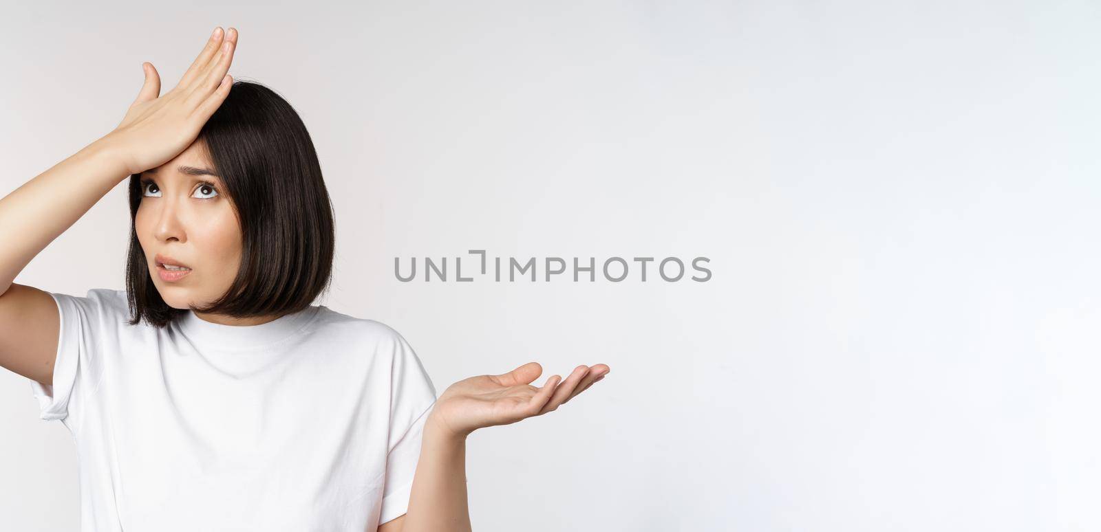 Annoyed korean girl facepalm, slap forehead and shrugging, confused by smth, standing over white background.