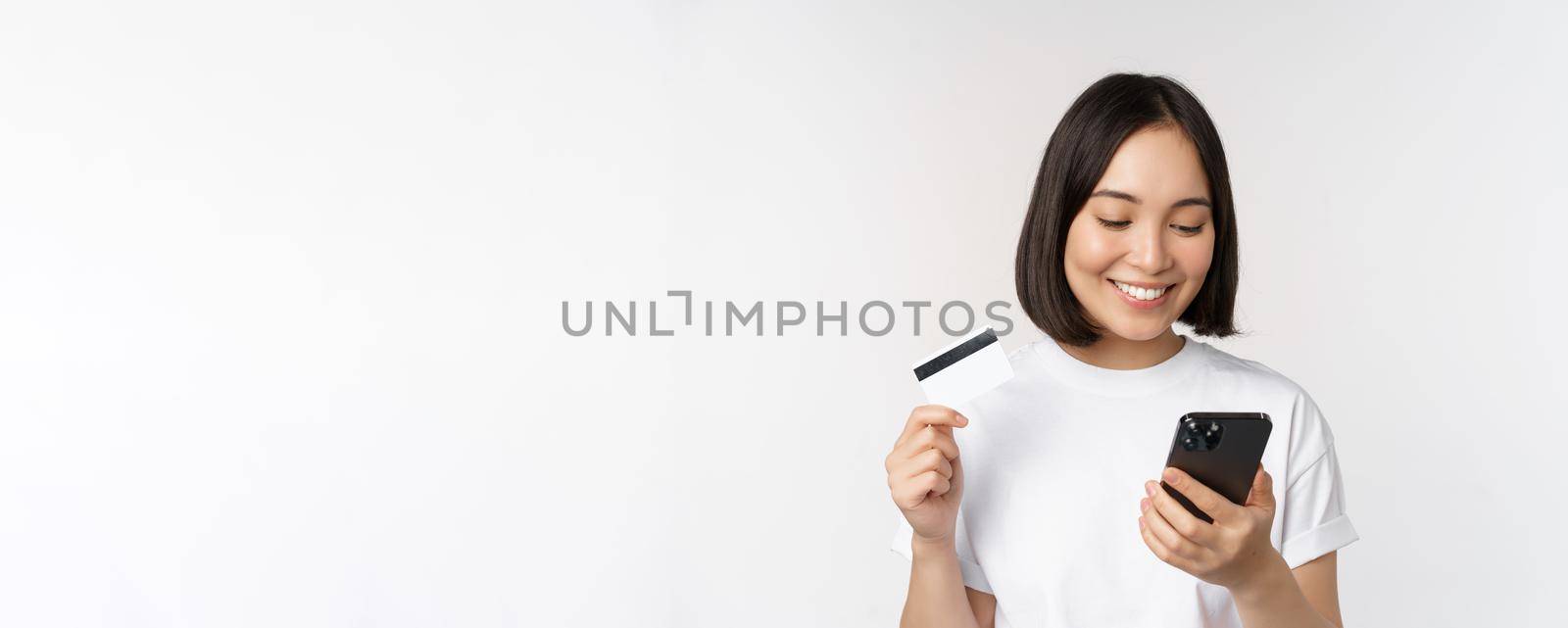 Online shopping. Happy asian woman using credit card and smartphone app, paying on website via mobile phone, white background by Benzoix