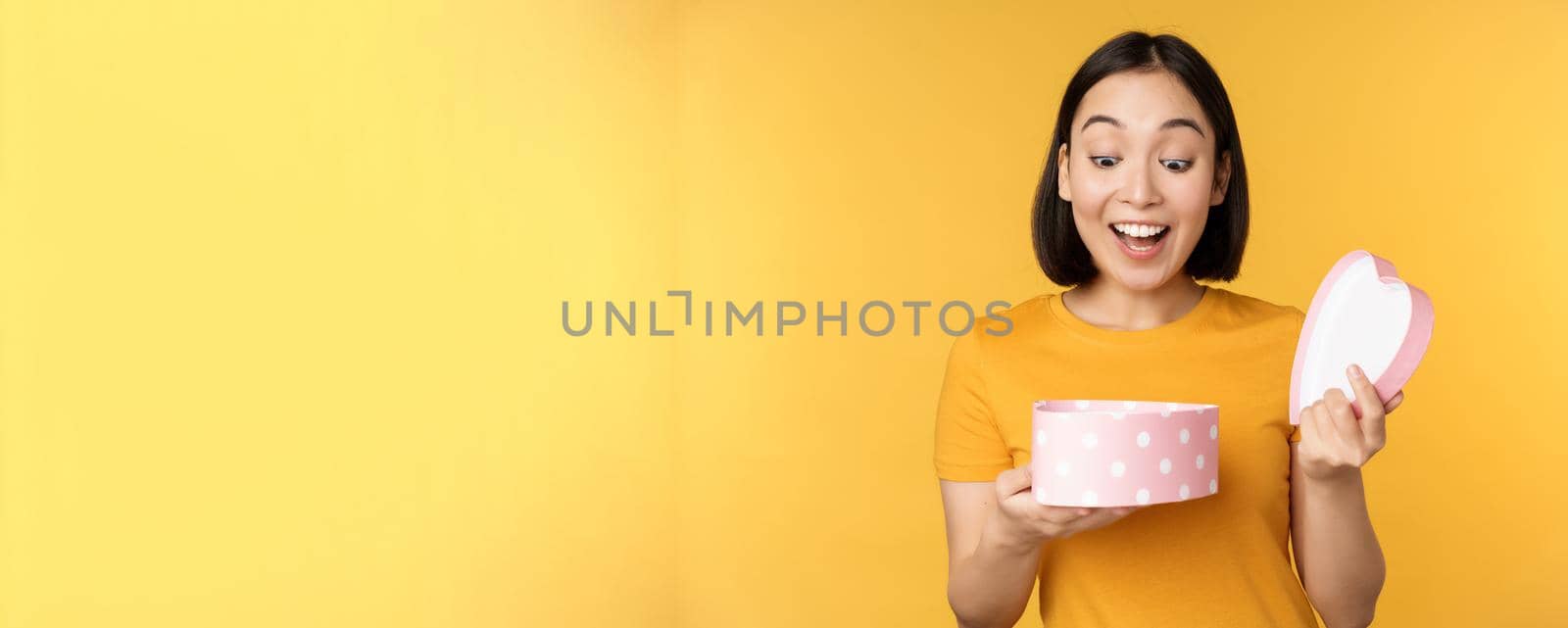 Portrait of excited asian woman, open gift box with surprised happy face, standing over yellow background.