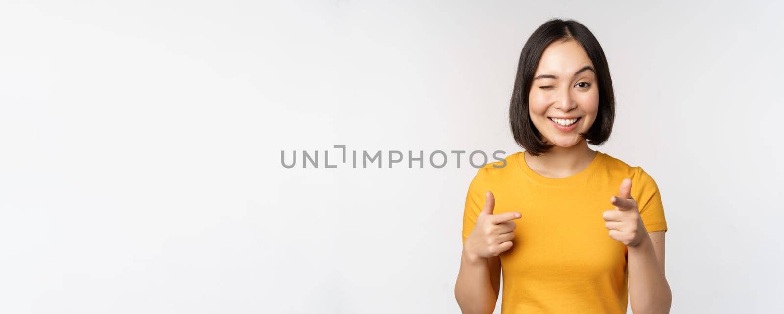 Young korean girl student, pointing fingers at camera and smiling, congratulating you, choosing, inviting people, standing over white background by Benzoix