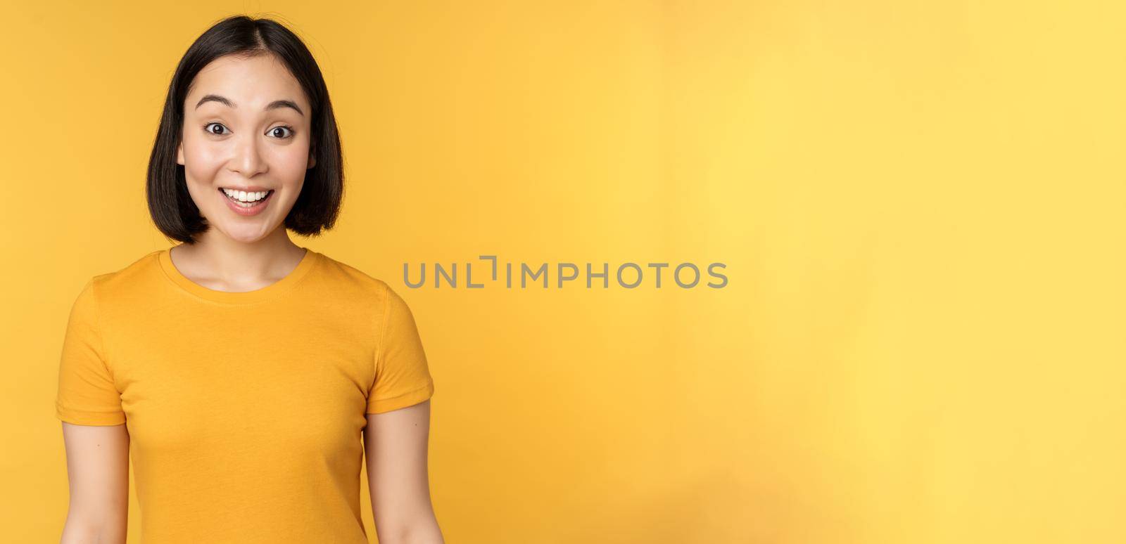 Image of asian girl looking surprised, reacting amazed, raising eyebrows impressed, standing over yellow background by Benzoix