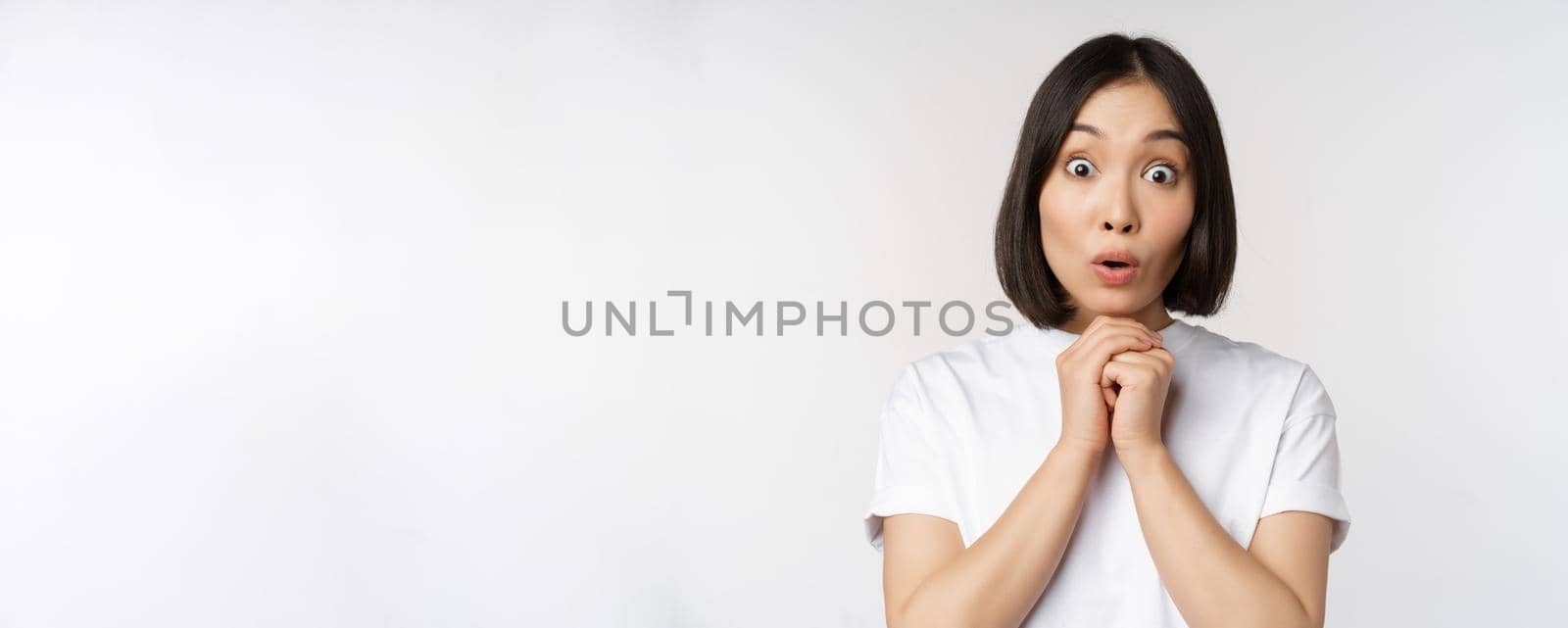 Close up portrait of asian brunette woman looking amazed, say wow, watching smth impressive, standing over white background by Benzoix