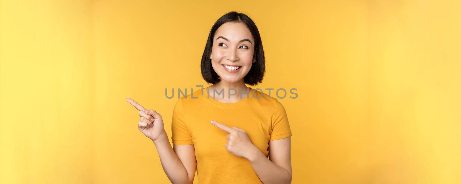 Portrait of happy asian girl pointing fingers and looking left, smiling amazed, checking out promo banner, showing advertisement against yellow background by Benzoix