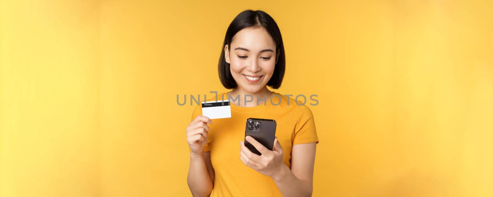 Online shopping. Smiling asian girl using credit card and mobile phone app, paying contactless, order on smartphone application, standing over yellow background.