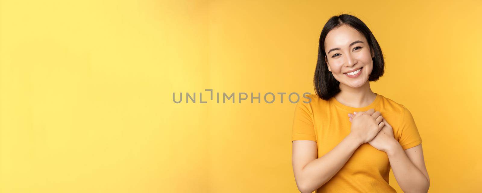 Romantic korean girl, asian woman holding hands on heart, smiling with care and tenderness, standing over yellow background.