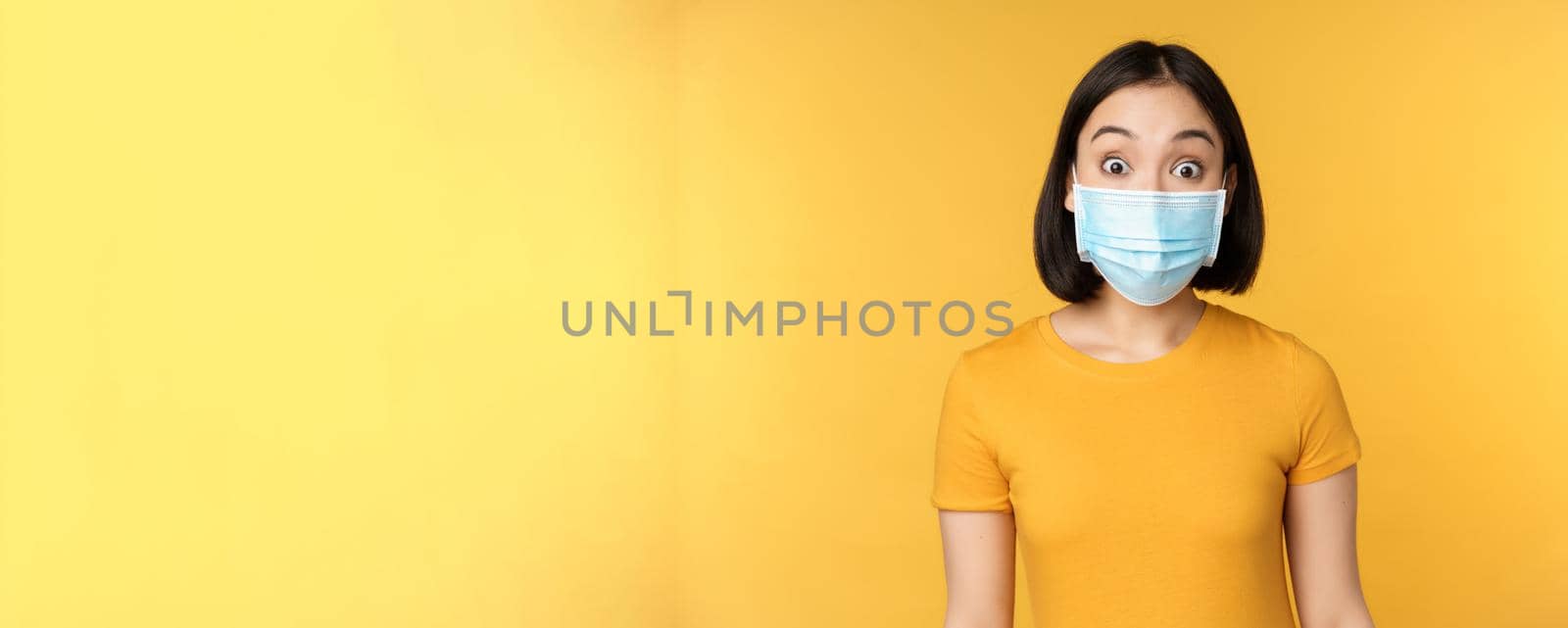 Portrait of korean girl in medical face mask looking surprised, amazed reaction to news, standing over yellow background.