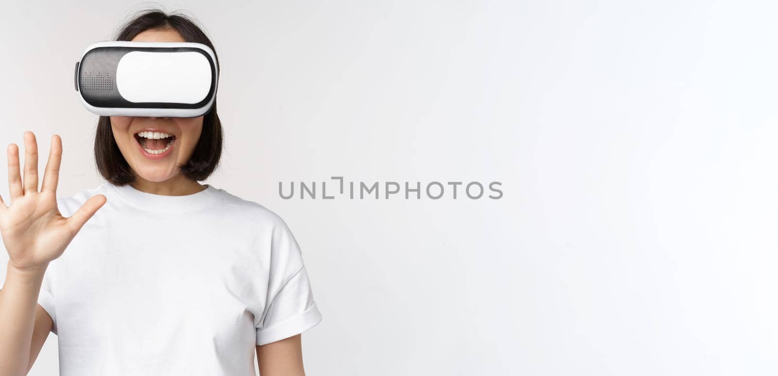 Happy asian woman using VR headset, waving raised hands and laughing, using virtual reality glasses, standing over white background.