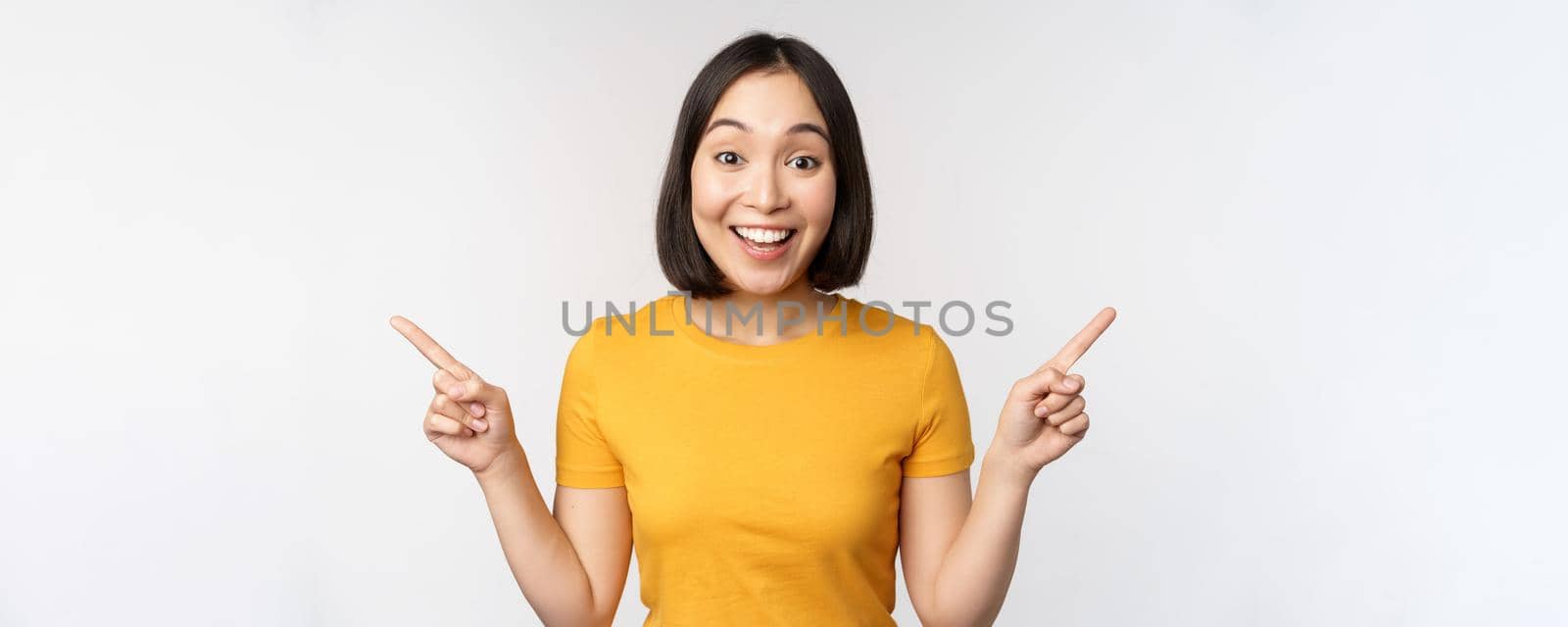 Cute asian girl pointing fingers sideways, showing left and right promo, two choices, variants of products, standing in yellow tshirt over white background by Benzoix