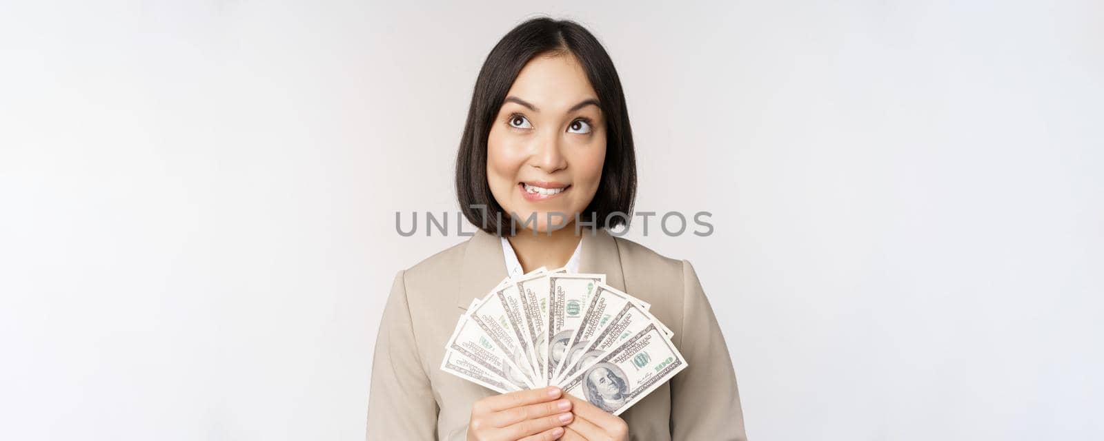 Image of asian corporate woman, happy businesswoman showing money, cash dollars and thinking, standing in suit over white background by Benzoix