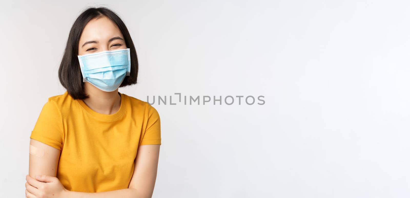 Healthy and happy asian woman in medical face mask, has band aid on shoulder after covid-19 vaccination, got vaccinated from omicron delta variant, whtie background.