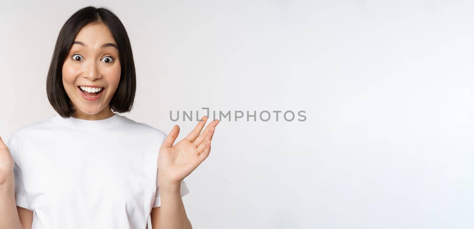 Close up portrait of asian girl looking surprised, wow face, reacting amazed at smth, standing in white tshirt over studio background, isolated.