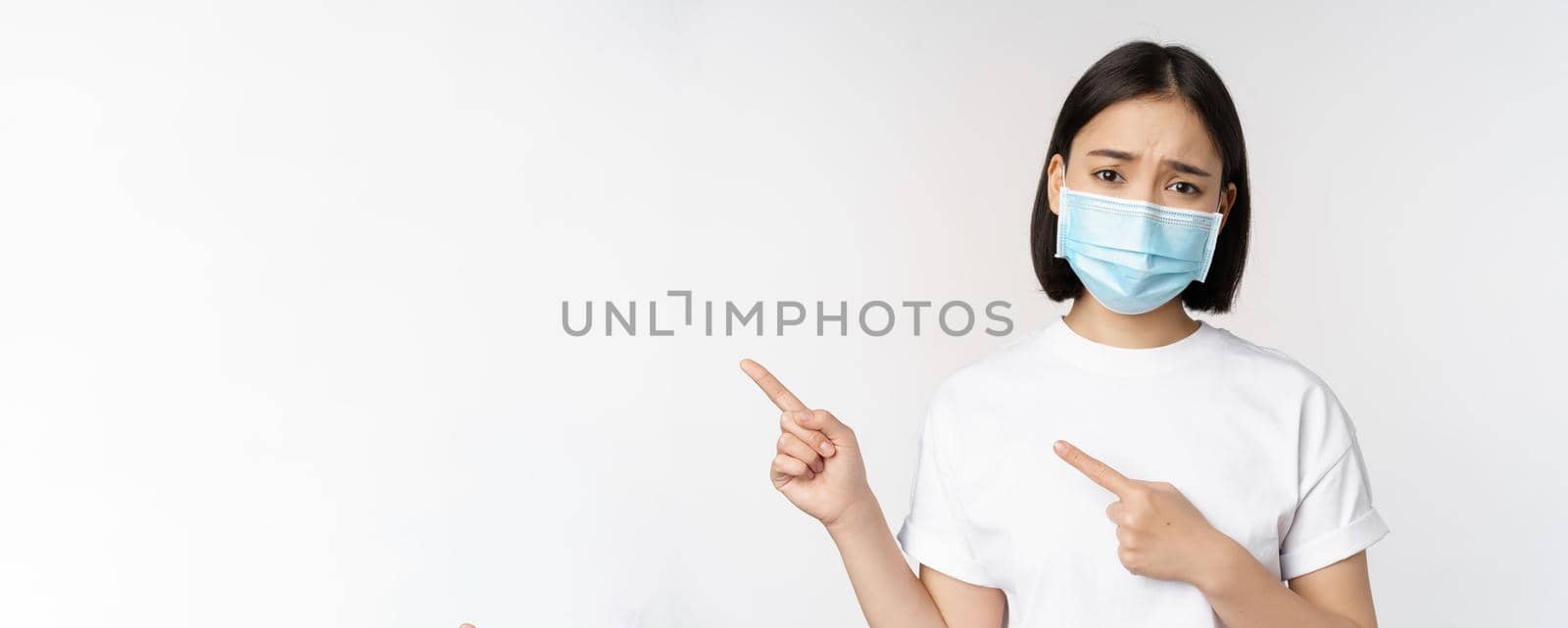 Sad asian woman in medical mask, pointing fingers left, frowning and looking upset, complaining, demonstrating banner, standing over white background by Benzoix