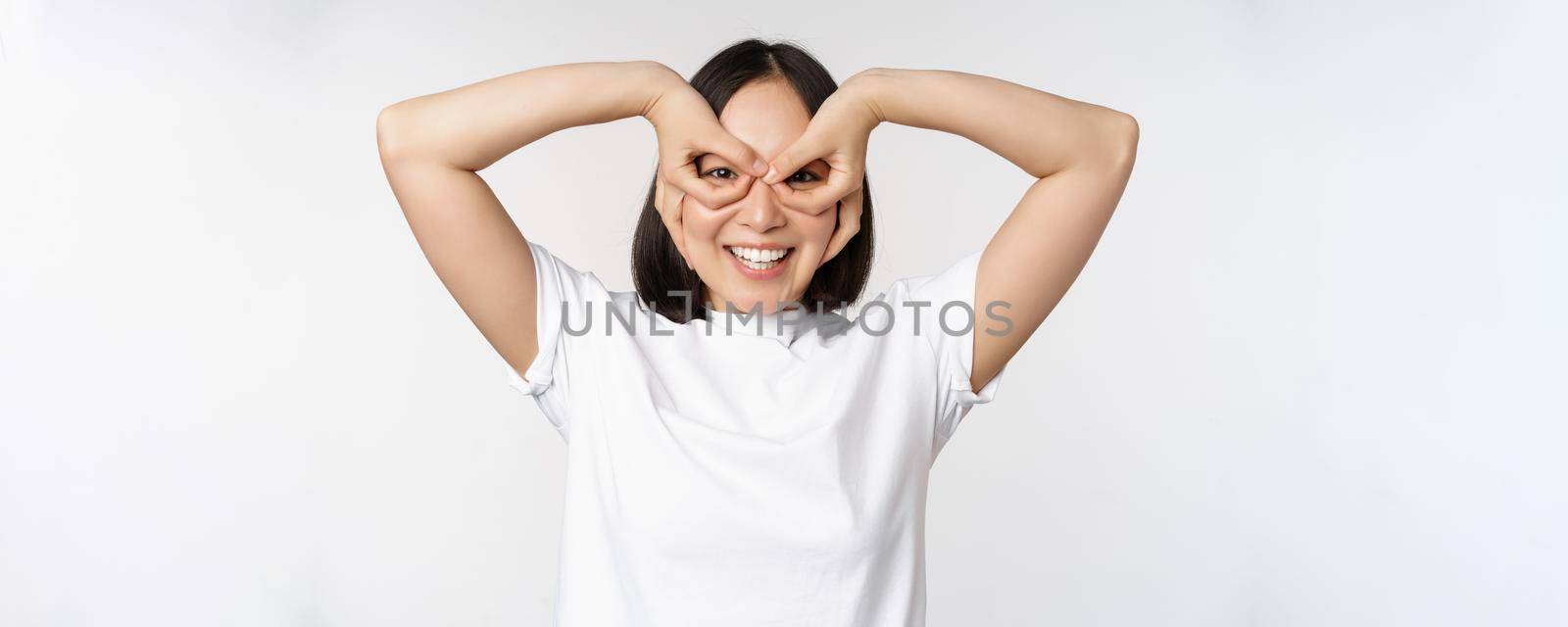 Funny young asian woman, korean girl making eyes glasses gesture, looking happy at camera, standing over white background by Benzoix