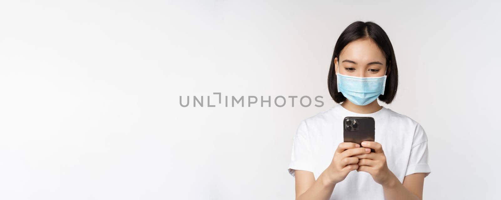 Health, covid and mobile concept. Young asian woman in medical face mask, looking at smartphone screen, using phone app, shopping online, white background by Benzoix