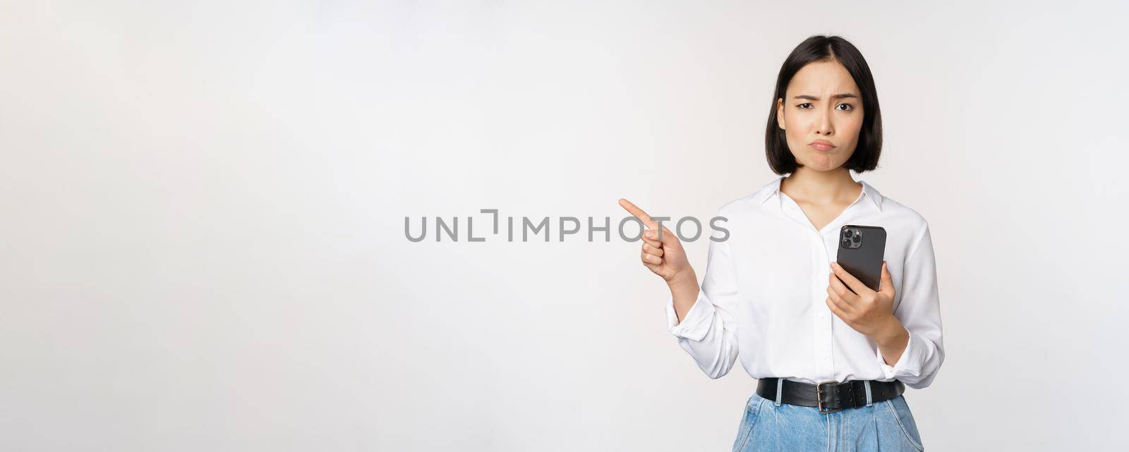 Skeptical asian modern girl, holding smartphone, pointing finger left at empty space and frowing hesitant, has doubts, standing over white background.