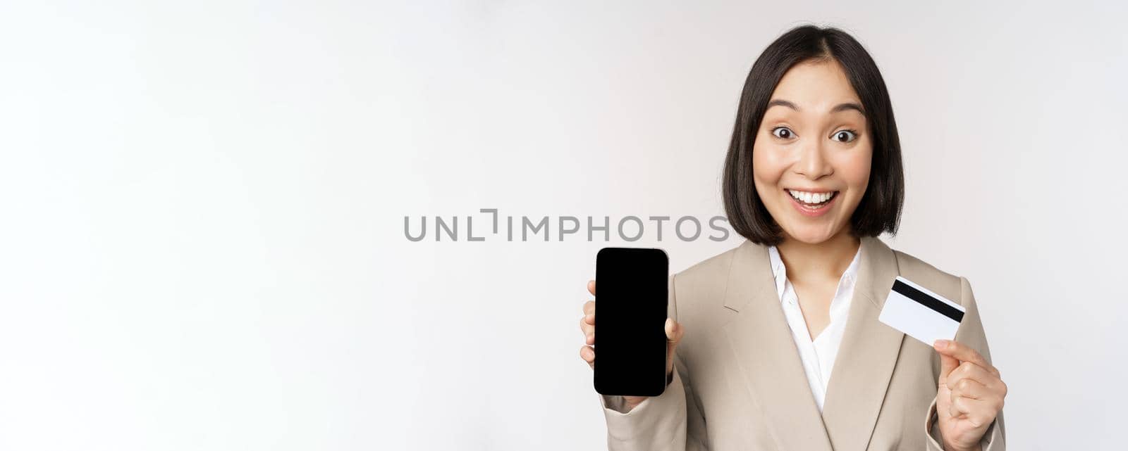 Corporate woman with happy, enthusiastic face, showing credit card and smartphone app screen, standing in suit over white background.